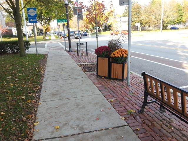 Trottoir de béton avec zone de commodité et d’agrément séparant la voie piétonnière de la voie de circulation routière. La zone de commodité et d’agrément est différenciée par un pavage de briques, ce qui produit un contraste de couleur et de texture. Un banc, des panneaux indicateurs et une boîte à fleurs y sont placés.