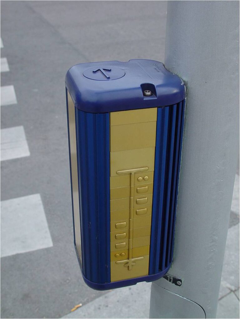 Photo of a pedestrian crossing with a push button that incorporates a tactile arrow to indicate the direction of crossing and a tactile map showing the lane configuration along the crossing route.